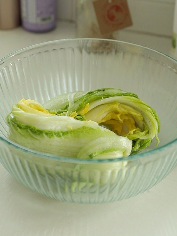 A glass bowl containing a halved head of napa cabbage with light green and yellow leaves, placed on a kitchen countertop.